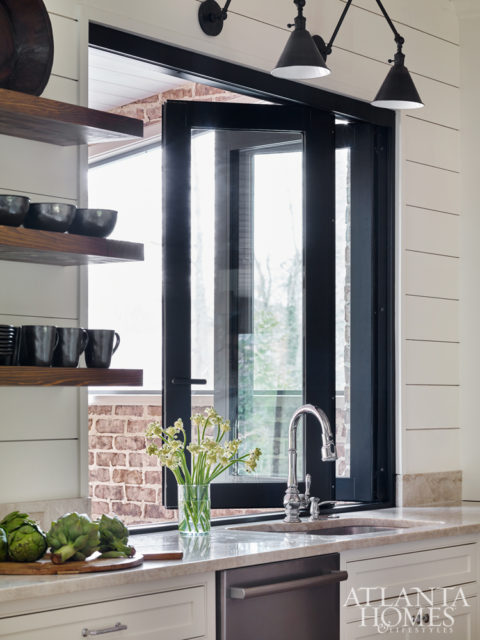 A kitchen window slides open as a pass-through to the outdoor dining space, next to open shelving with everyday dinnerware.