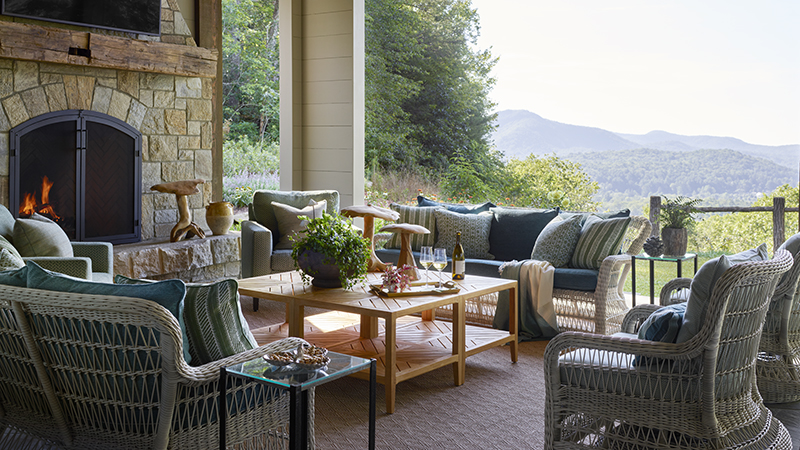 Read article: Inside a Colorful Cashiers Mountain Retreat Designed for Modern Family Living Covered porch with stone fireplace, wicker seating, and mountain view