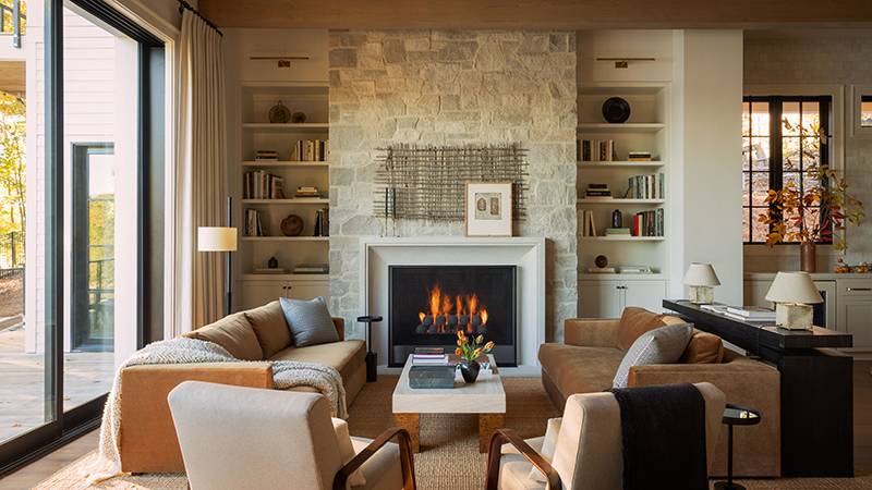 Read article: How Brian Cash Works and MAK + KAM Created a Modern Lake Lanier Retreat That Feels Both Grand and Inviting Living room with stone fireplace, built-in bookshelves, and tan sofas.