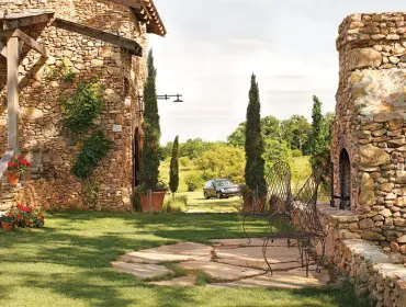 Stone house with arched doorways and wooden beams, featuring a stone fireplace, wrought iron chairs, and cypress trees in a grassy yard with a car in the distance.
