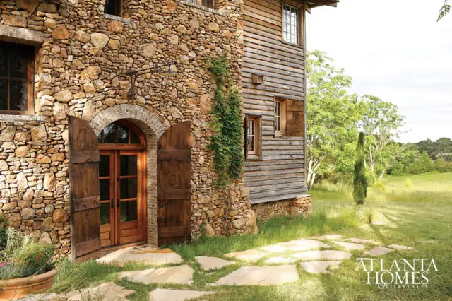 Stone and wood exterior of a rustic home with arched double doors, wooden shutters, and a flagstone path leading to the entrance. Lush green landscape surrounds the building.