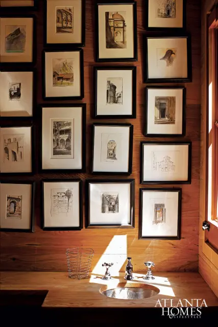 Wall of framed architectural sketches and watercolors above a wooden countertop with a metal sink and chrome faucets.