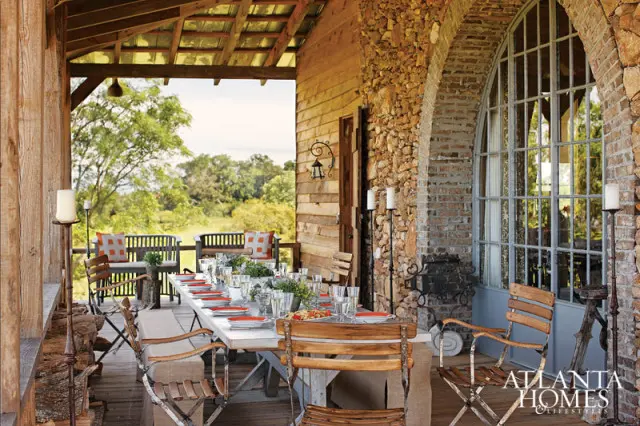 Covered porch with a long dining table set for a meal, rustic wood siding, stone accents, and a large arched window.
