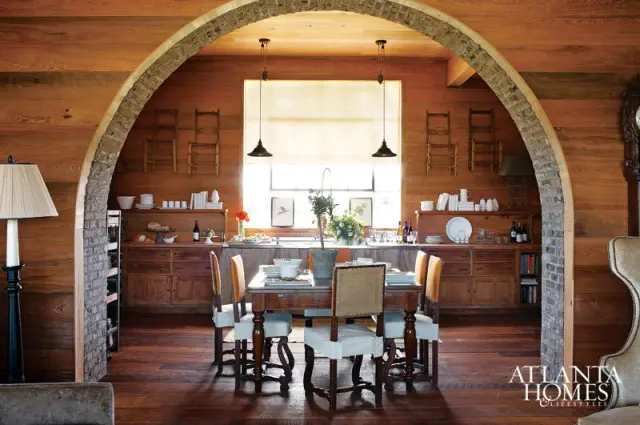 Rustic dining room with wood-paneled walls, a large wooden table set for a meal, and a stone archway leading to a kitchen area with a sink and cabinetry.