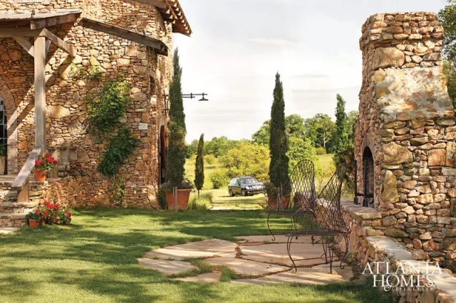 Stone house with arched doorway, outdoor fireplace, and wrought iron chairs on a flagstone patio with a manicured lawn and rolling hills in the background.