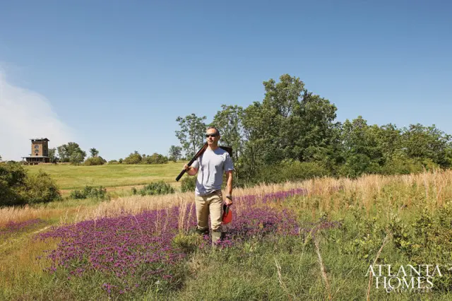 Man carrying a shotgun walks through a field of purple flowers towards a tower in the distance under a clear blue sky.