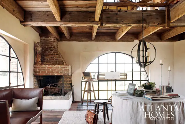 Rustic living room with exposed wooden beams, a large brick fireplace, arched windows, and a round table covered with a linen cloth displaying books and plants.