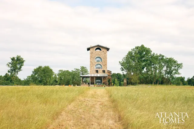 Stone tower with arched windows and a covered porch, set in a grassy field with trees under a cloudy sky. Atlanta Homes & Lifestyles logo in the lower right corner.