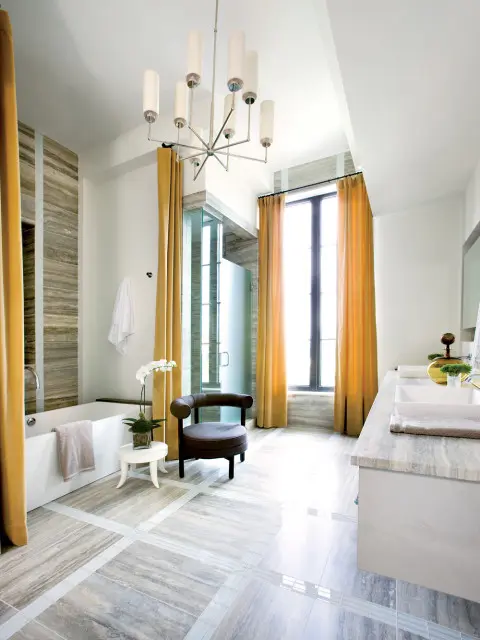 Modern bathroom with a freestanding tub, glass-enclosed shower, double vanity with marble countertop, and a dark upholstered chair. Ochre curtains frame a tall window.