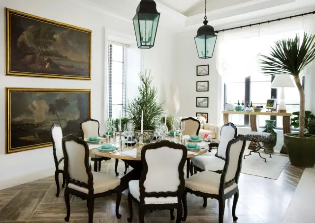 Dining room with round table set for eight, featuring ornate dark wood chairs with cream upholstery, large landscape paintings on the wall, and hanging lantern lights.