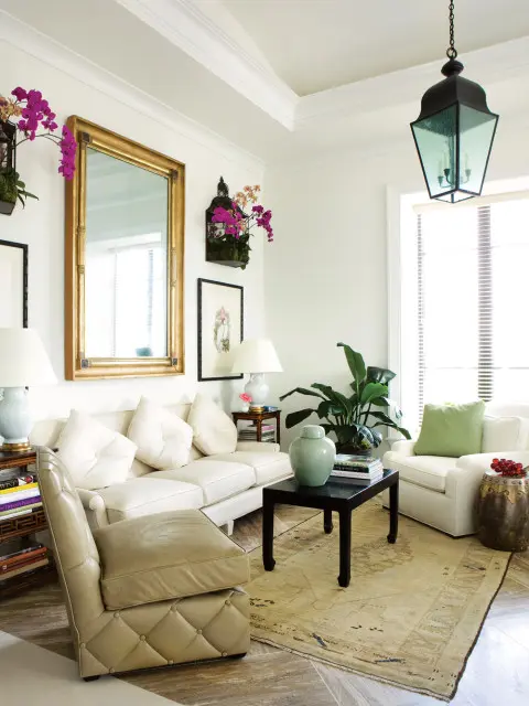 Cream sofa and armchair in a living room with a large gold-framed mirror, orchids, and a dark wood coffee table with a jade vase.