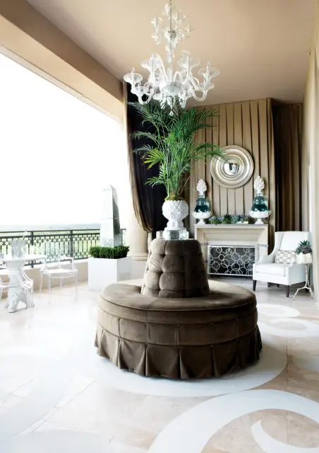 Elegant sunroom with a tufted circular sofa, a glass chandelier, a fireplace, and clear acrylic chairs on a balcony overlooking greenery.