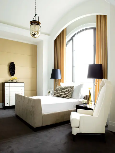 Bedroom with a beige upholstered bed, white armchair, black lamps, and a large arched window with gold curtains. A gold textured wall is behind a black and white dresser.