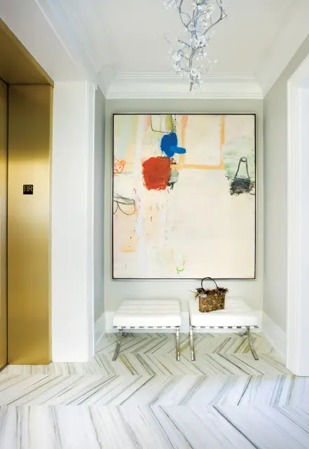Hallway with gold elevator doors, abstract art, two white tufted benches, and marble herringbone flooring.
