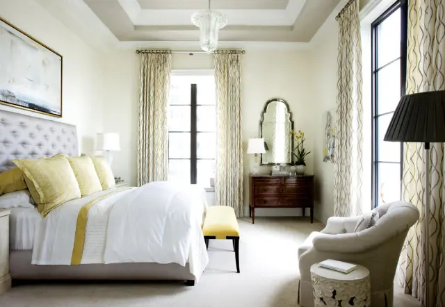 Bedroom with a tufted gray headboard, yellow accent pillows, white bedding, and patterned curtains. A dark wood dresser with a mirror sits opposite the bed.