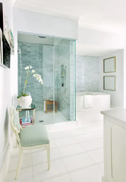 Light blue and white bathroom with glass-enclosed shower with marble tile, a freestanding tub, and a decorative chair.