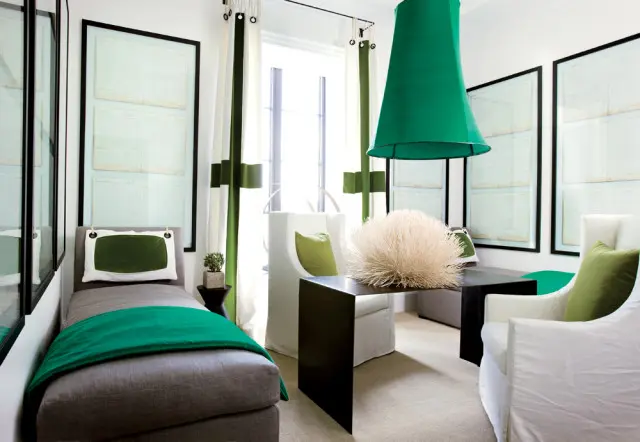 Gray chaise lounge with emerald green throw and pillow, white armchair, and large green pendant light in a room with framed documents on the wall.