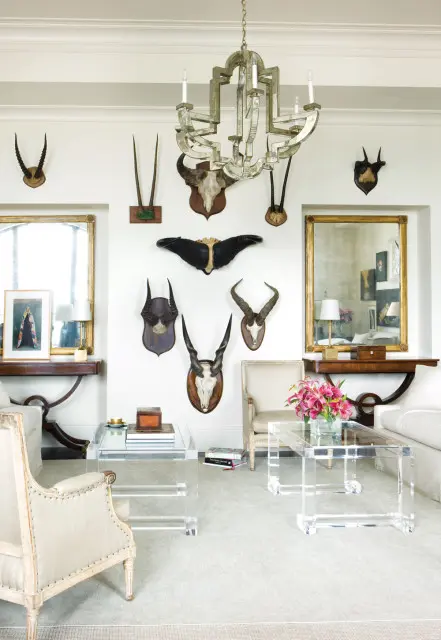 Living room with white walls featuring taxidermied animal heads and horns, a mirrored chandelier, two acrylic coffee tables, and upholstered chairs.