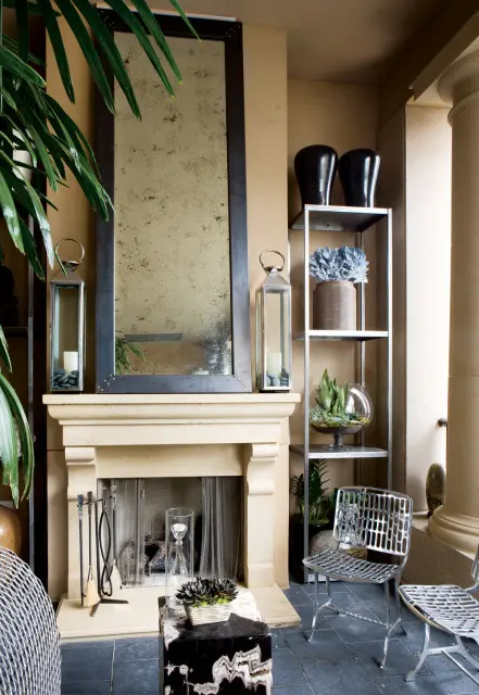 Living room with a light stone fireplace, large distressed mirror, metal shelving unit with decorative items, and two silver chairs.