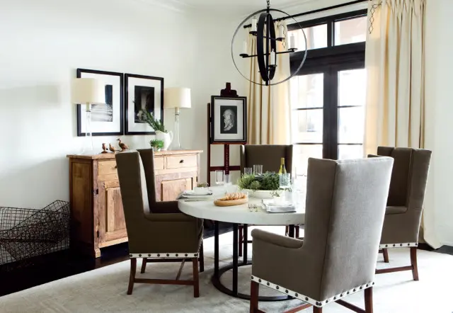 Dining room with round concrete table, upholstered chairs with nailhead trim, antique wood buffet, and black circular chandelier.