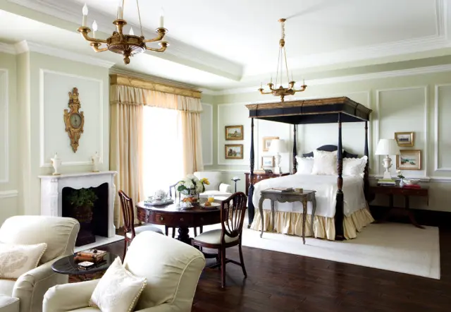 Light green bedroom with a dark wood four-poster bed, white bedding, ornate fireplace, round dining table with breakfast setting, and plush armchairs.