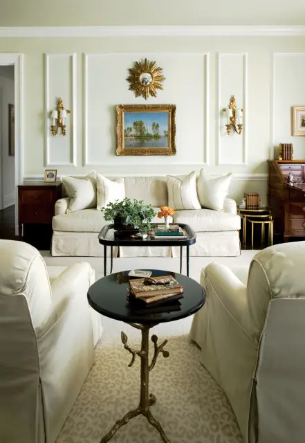 Pale green living room with a cream sofa, two armchairs, and a black coffee table with books and plants. A landscape painting hangs above the sofa.