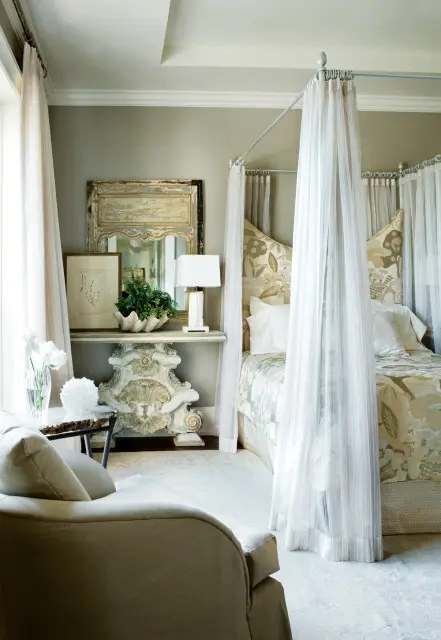 Bedroom with a four-poster bed draped in sheer white fabric, a floral headboard, and a chaise lounge in the foreground. A console table with a distressed mirror and lamp sits against the wall.