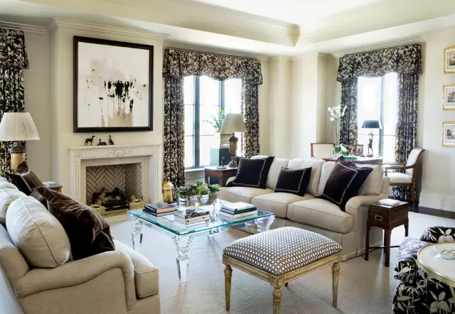Living room with a neutral-toned sofa, dark patterned curtains, a glass coffee table, and a fireplace with abstract art above.