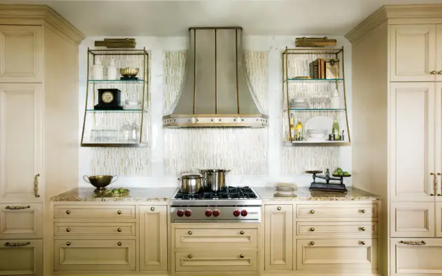 Cream kitchen with a stainless steel range hood, marble backsplash, and glass-fronted shelves displaying decorative items and dishware.