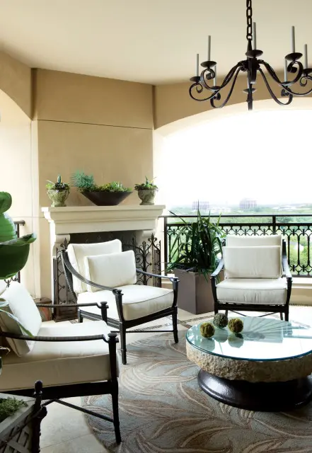 Covered balcony with cream upholstered chairs, a stone and glass coffee table, a fireplace, and a wrought iron chandelier.