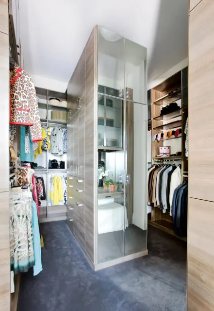Walk-in closet with grey carpet, wood-grain cabinetry, mirrored doors, and clothing racks filled with garments and accessories.