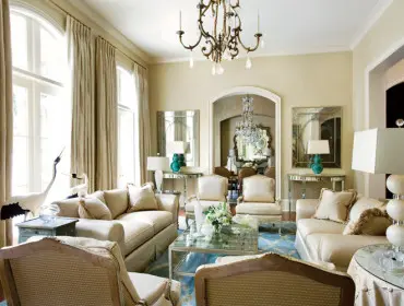 Living room with cream sofas, armchairs, and a glass coffee table. Tall arched windows are draped in cream curtains. A chandelier hangs overhead, and a dining room is visible through an archway.