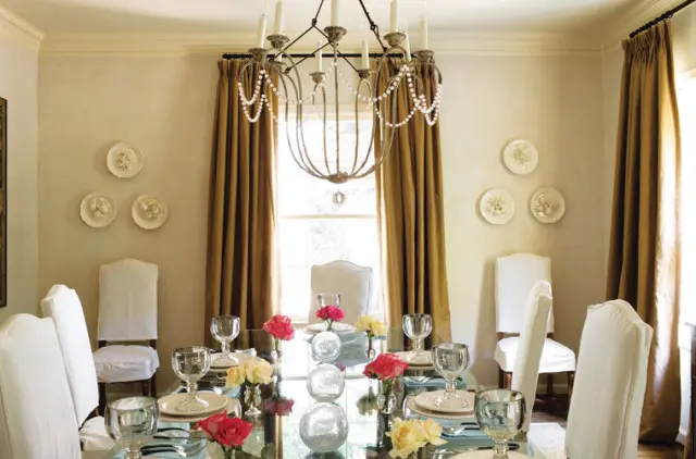 Dining room with a glass-top table set for a meal, white slipcovered chairs, gold drapes, and decorative plates on a neutral wall.