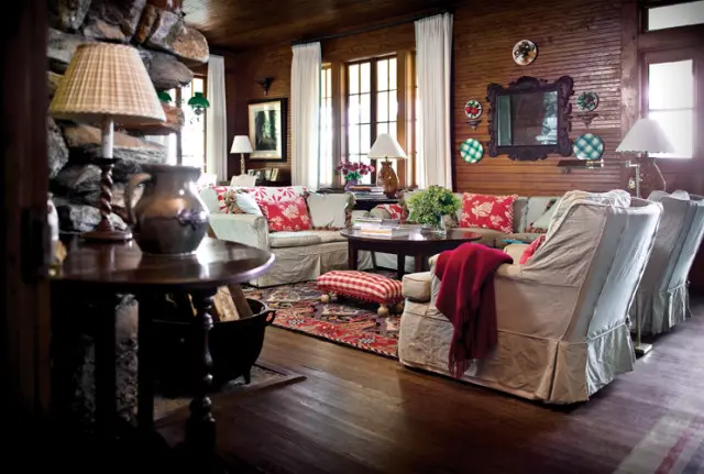 Living room with stone fireplace, wood-paneled walls, slipcovered sofas with red floral pillows, and a patterned rug.