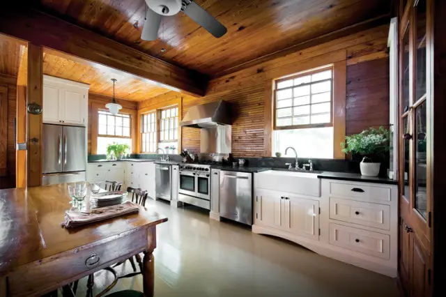 Kitchen with white cabinetry, stainless steel appliances, farmhouse sink, and wood-paneled walls and ceiling. A wooden dining table is in the foreground.