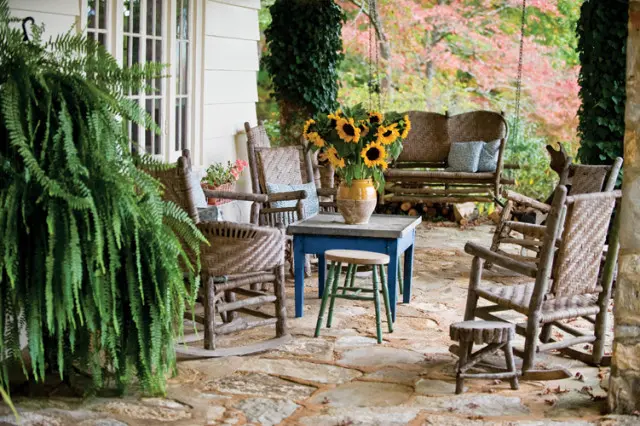 Porch with stone flooring, wicker rocking chairs, a blue table with sunflowers, and a wooden swing.
