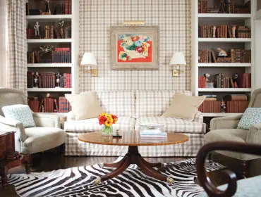 Living room with beige and white gingham wallpaper, two armchairs, a sofa, built-in bookshelves filled with books, and a round wooden coffee table on a zebra print rug.