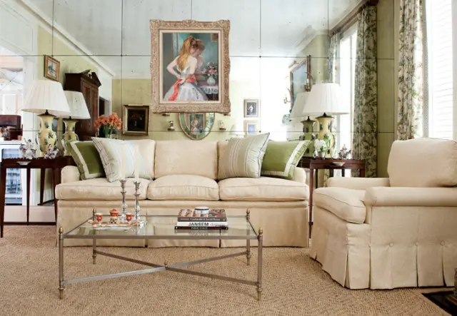 Cream sofa and armchair with green and striped pillows in a living room with mirrored walls, a glass coffee table, and floral curtains.