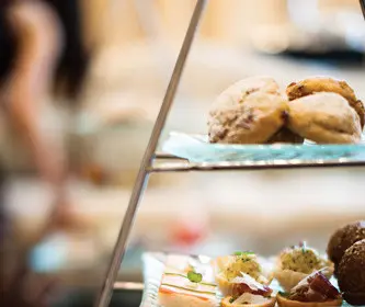 Tiered serving tray with assorted pastries, including scones, small sandwiches, red and white layered jelly, gingerbread cookies, and chocolate desserts.