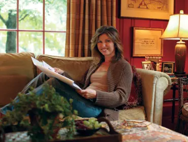 Woman sitting on a couch holding papers, wearing a brown cardigan and jeans. A window with green trees is visible behind her.