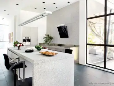Modern white kitchen with a large speckled granite island, stainless steel appliances, and a black range hood mounted on a textured wall.