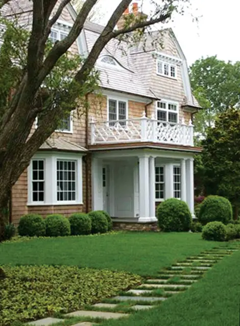 Shingle-sided house with a gambrel roof, white columns, a balcony with lattice railing, and a stone pathway leading to the front door.