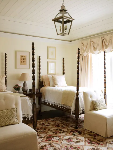 Bedroom with two four-poster beds, cream walls, wood floors, and a patterned rug. A large lantern hangs from the paneled ceiling.