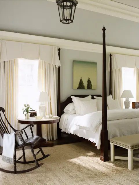 Bedroom with a dark wood four-poster bed, white bedding, a wooden rocking chair, and a round side table with lamps.