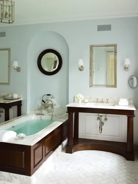 Light blue bathroom with a marble-clad soaking tub, dark wood vanity with marble countertop, and herringbone marble tile flooring.
