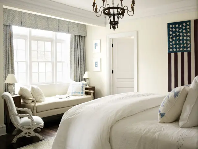 Cream bedroom with a large window dressed in blue floral curtains, a cream daybed with pillows, a white upholstered chair, and an American flag tapestry on the wall.