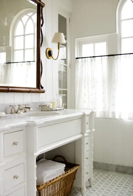 White bathroom vanity with marble countertop, silver fixtures, and a wicker basket with folded white towels. Paneled walls and a patterned tile floor.