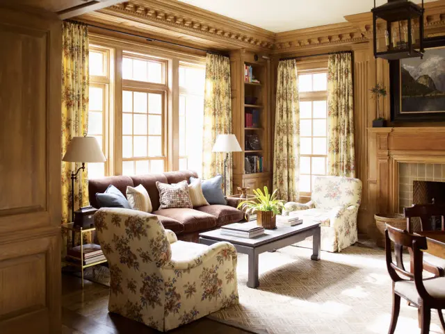 Living room with wood paneled walls, floral print armchairs, a brown sofa, and patterned curtains. A dark wood coffee table sits in the center of the room.