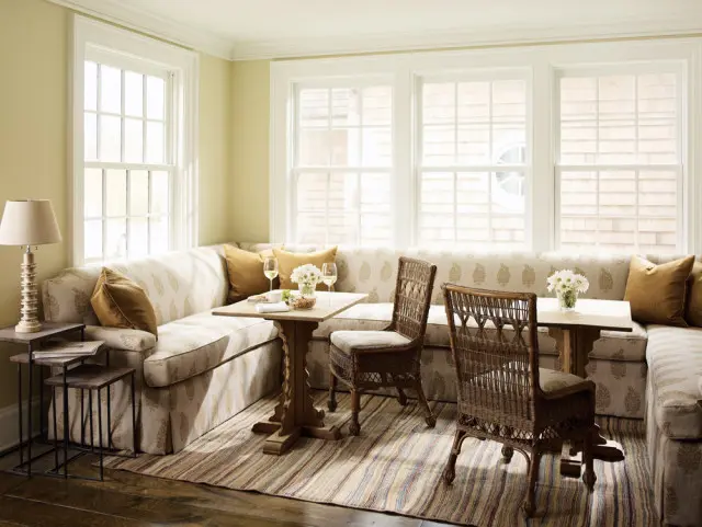 Breakfast nook with a built-in sectional sofa upholstered in a cream and gold floral pattern, two wicker chairs, and a wooden pedestal table.