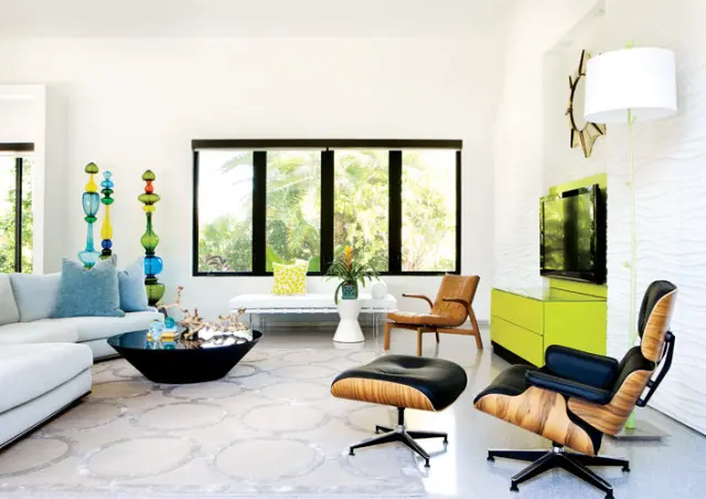 Living room with white sectional sofa, black coffee table, Eames lounge chair and ottoman, and a lime green media console. Large windows overlook a lush green landscape.
